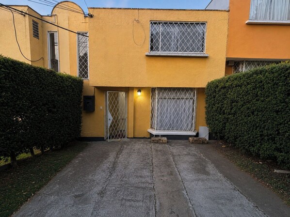 A two-story building with a gated entrance, a small window with a metal grille, and a light fixture above the door. - Jardines del Bosque – Your Quito Getaway (Pichincha)