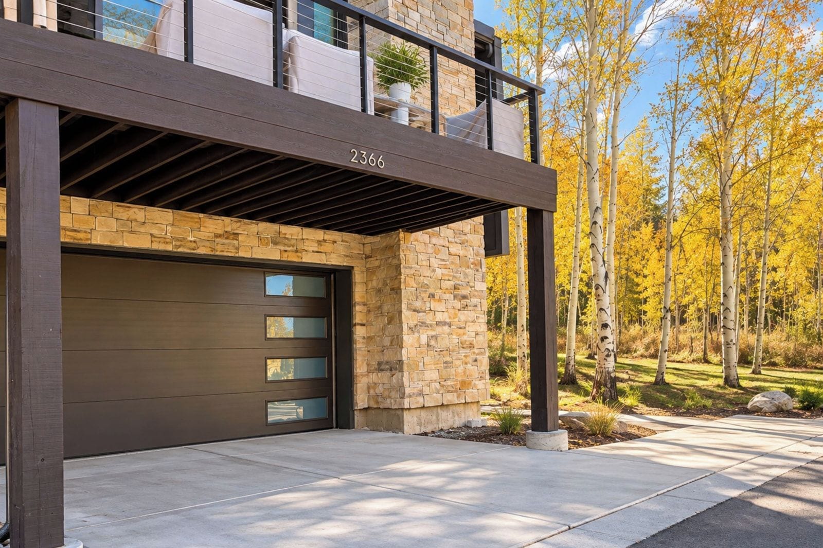 A modern two-story house with a garage, stone facade, and a balcony overlooking a landscaped yard with trees.