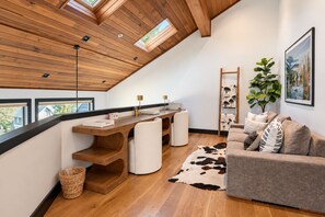 A modern loft-style room with a sloped ceiling, wooden beams, a dining area with a white table and chairs, a sofa with patterned cushions, and a large window offering a view of the outdoors. - Queen Esther Townhome by KBM (Park City)