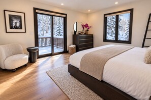 A bedroom with a large bed, a chair, a dresser, and a view of snowy trees through the windows. - Queen Esther Townhome by KBM (Park City)