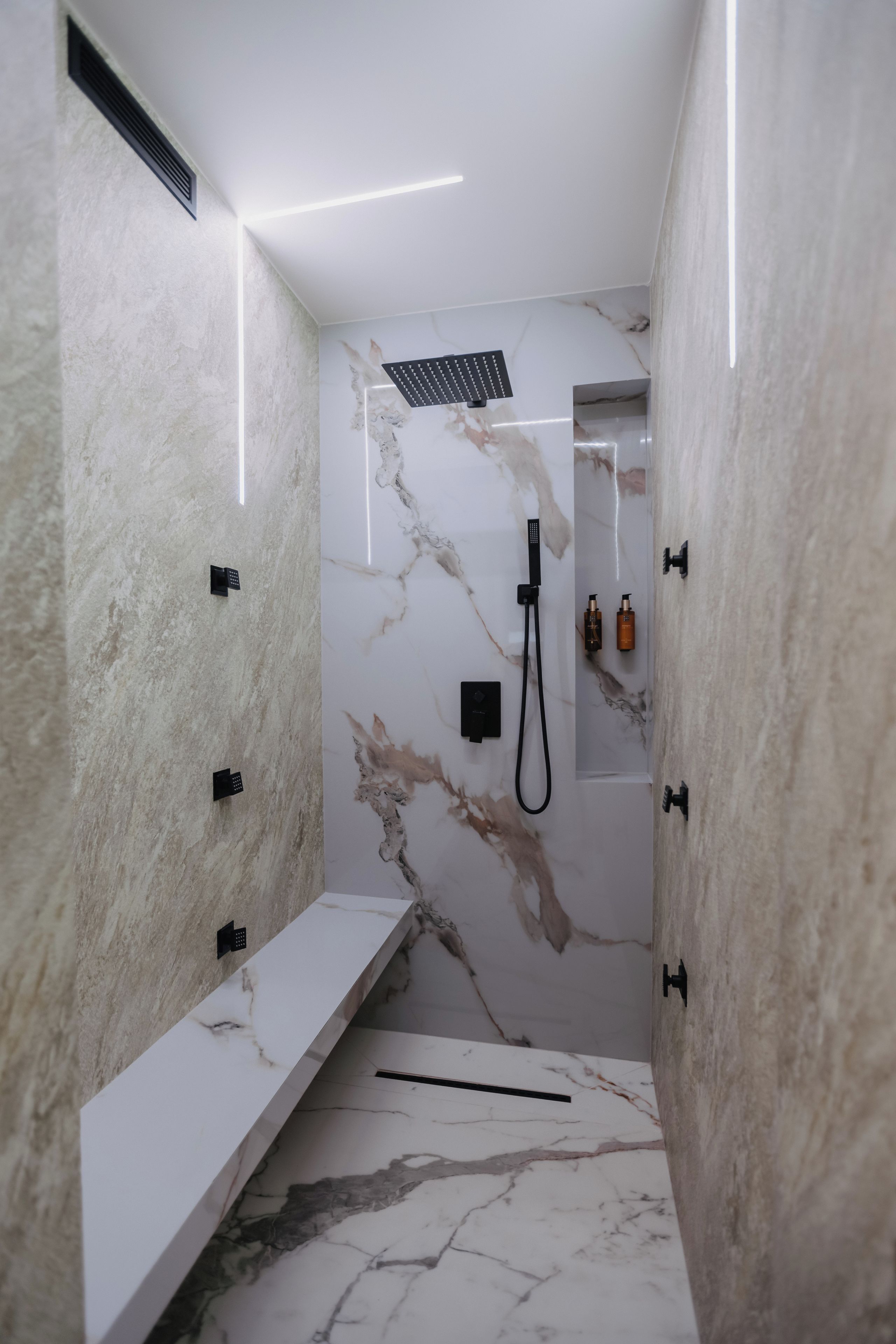 A modern bathroom with a marble wall, a shower with a handheld showerhead, and a shelf with toiletries.