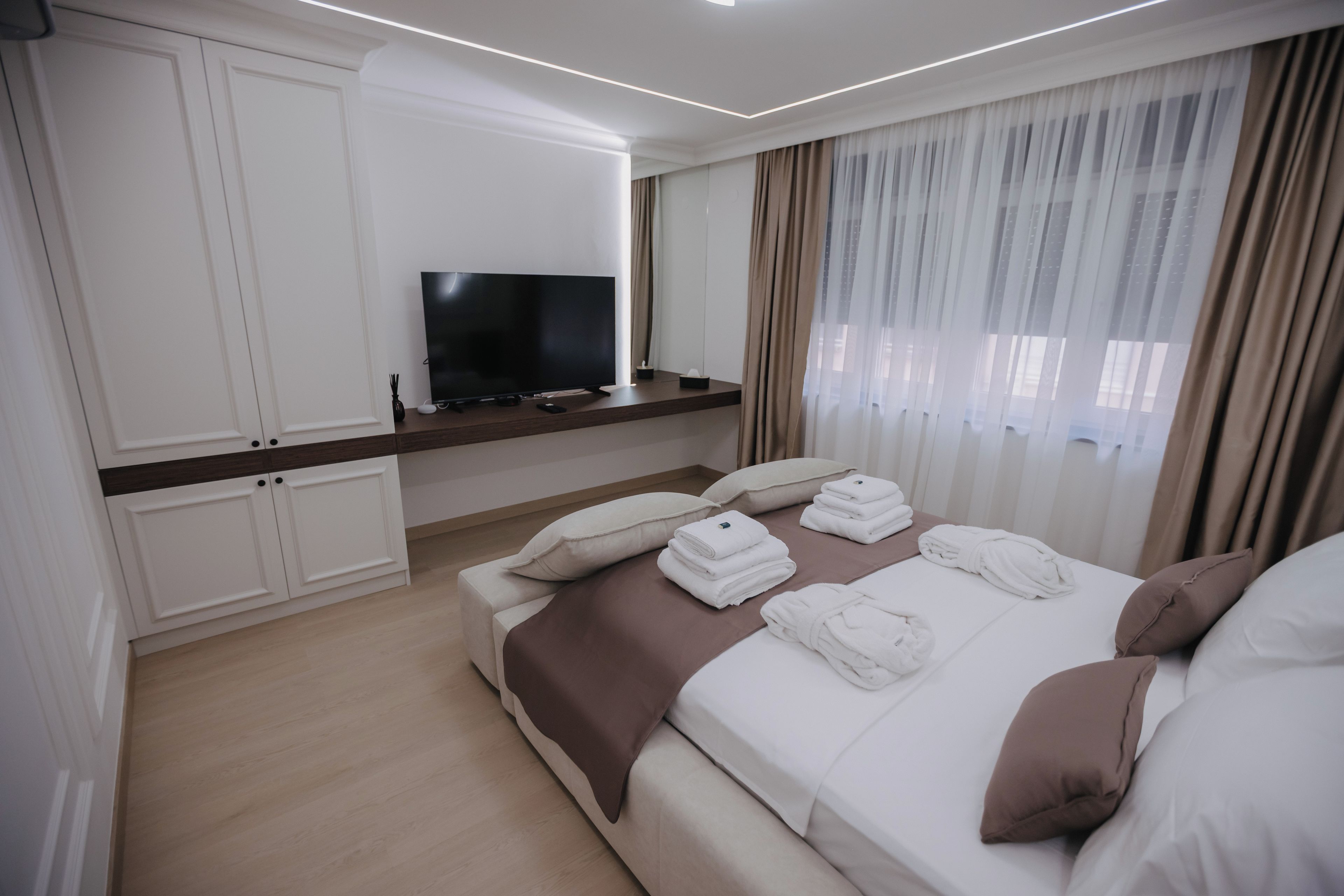 A modern hotel room with a flat-screen TV mounted on a wall, a bed with neatly folded towels, and a large window with sheer curtains.