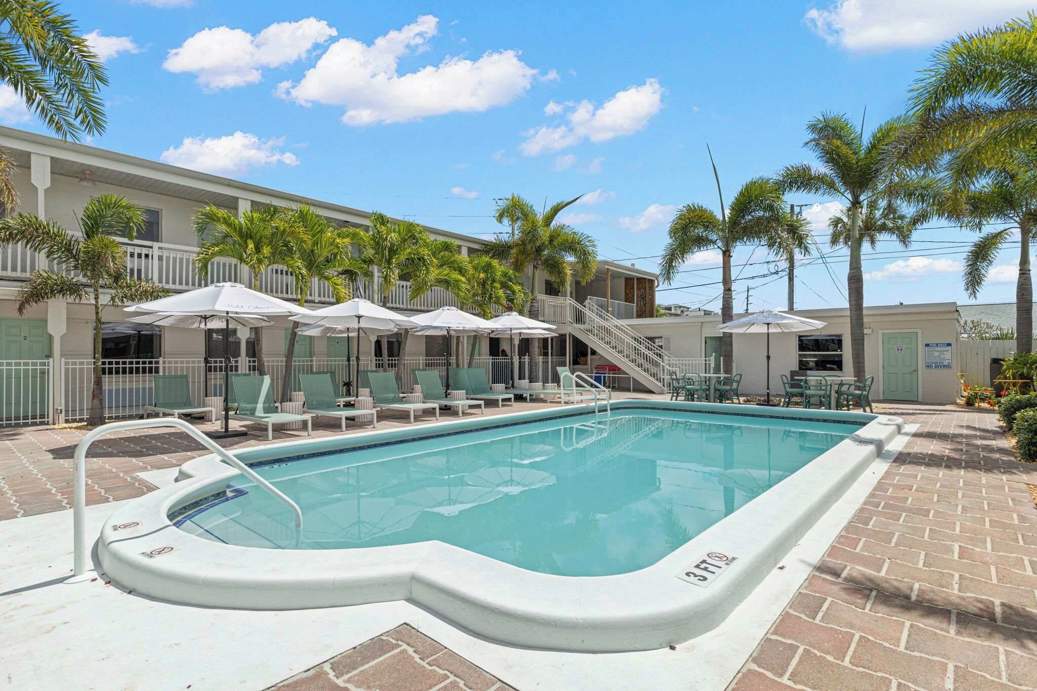 A swimming pool with lounge chairs and umbrellas, surrounded by palm trees and a building with a balcony.