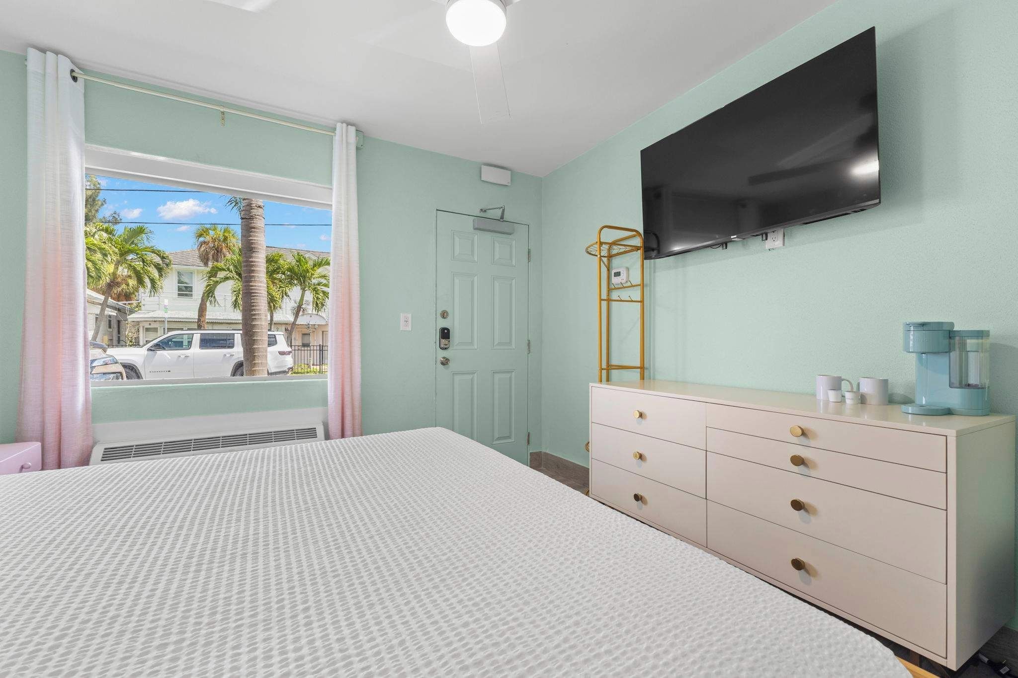 A bedroom with a bed, dresser, TV, and a window with a view of palm trees and a car.