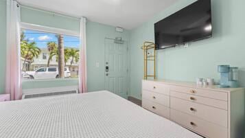 A bedroom with a bed, dresser, TV, and a window with a view of palm trees and a car.
