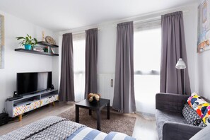 Ein modernes Wohnzimmer mit Flachbildfernseher, grauen Vorhängen, einer Couch und einem gemusterten Teppich.