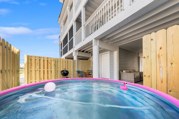 Une piscine avec un bord rose, un flotteur en forme de flamant et un barbecue dans une cour arrière agrémentée de clôtures en bois et d’un patio couvert.