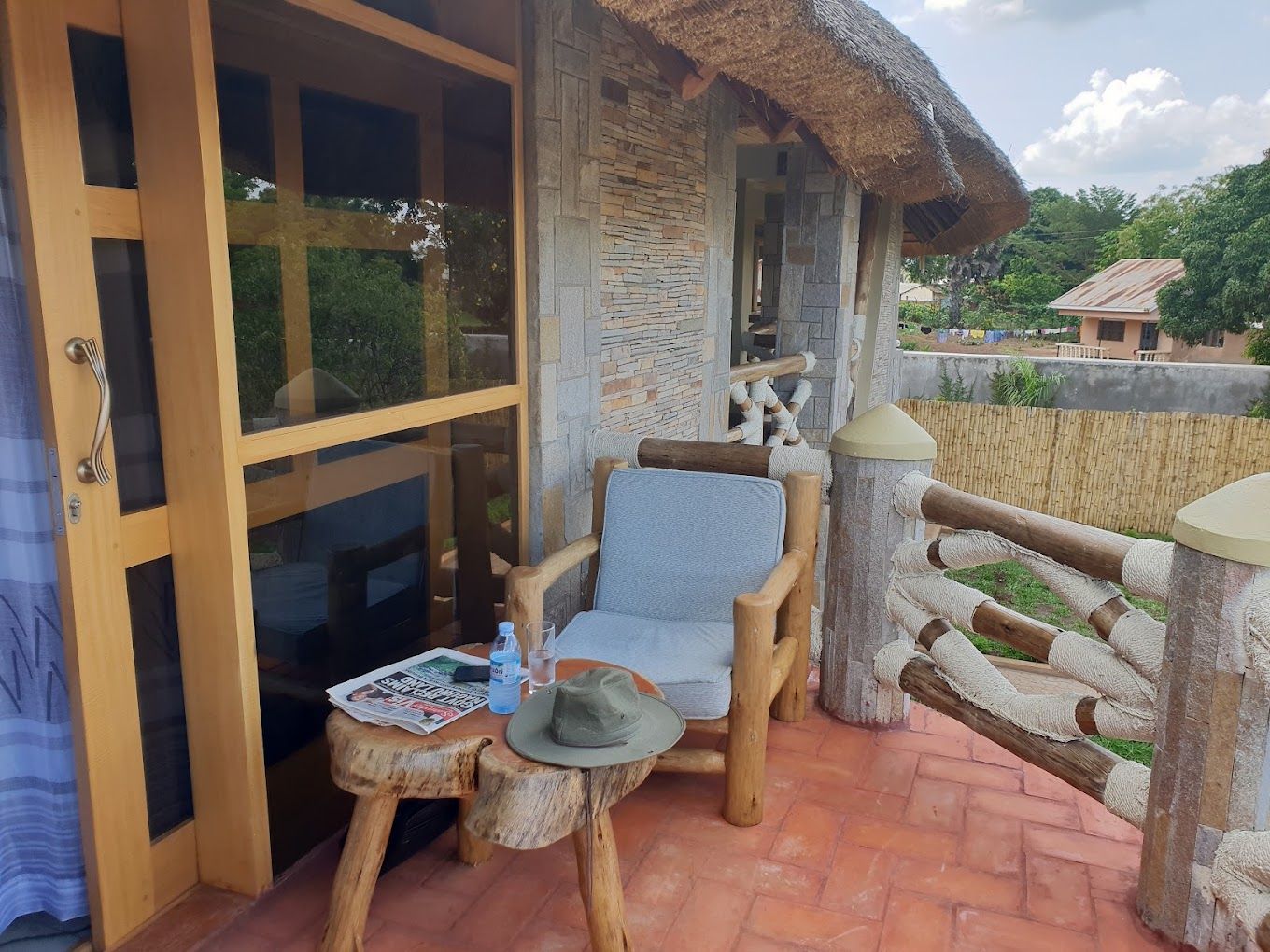 A wooden deck with a chair and a small table, a glass door, and a stone wall with a thatched roof.