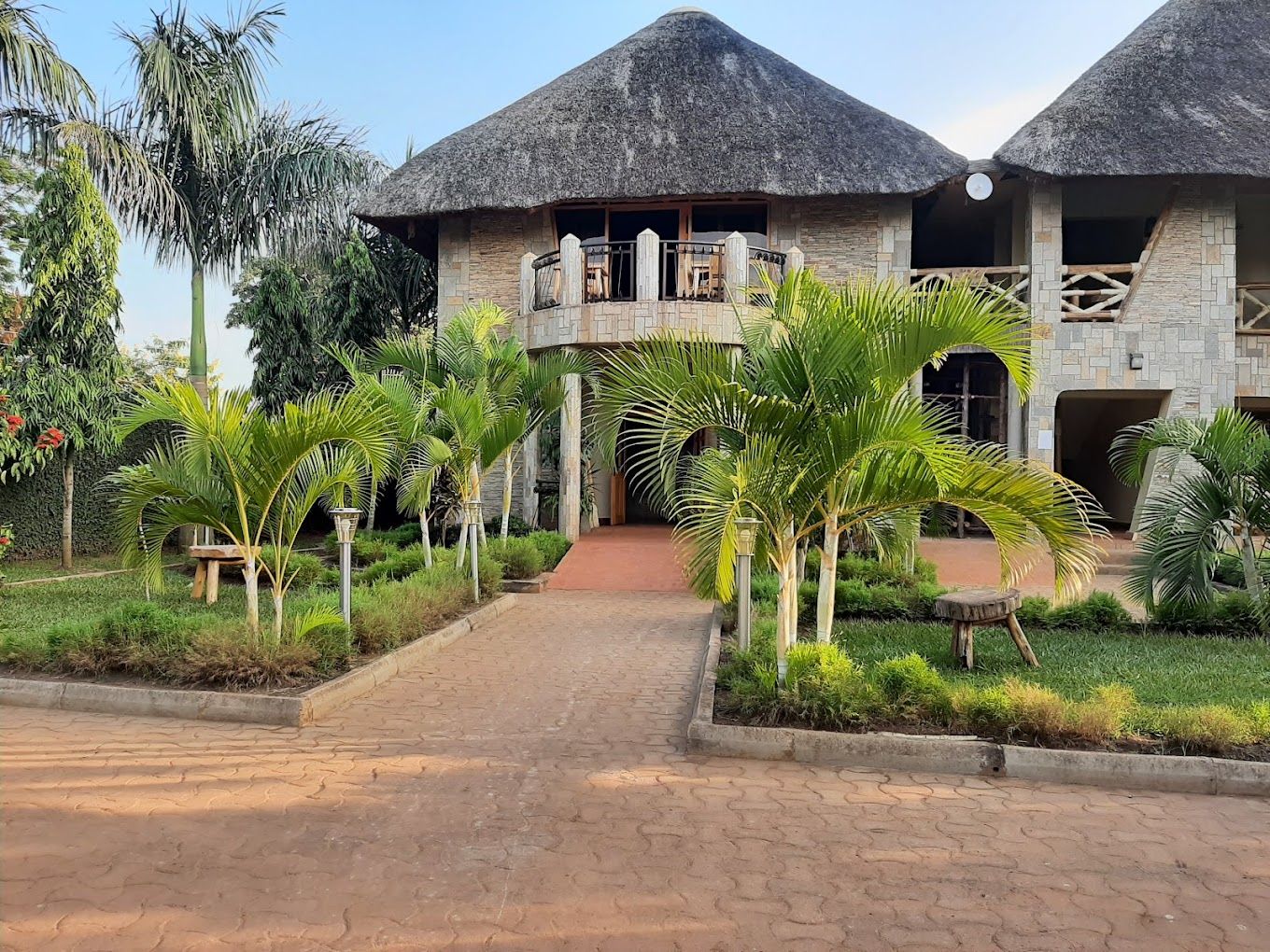 A building with a thatched roof and stone walls, a paved entrance, and a garden with palm trees.