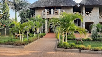 A building with a thatched roof and stone walls, a paved entrance, and a garden with palm trees.