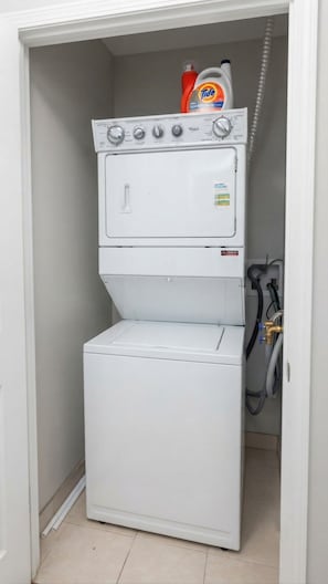 A compact laundry room with a stacked washer and dryer, a hose, and a detergent bottle. - Chic Mississauga Condo with Pool, Gym & City Views (Mississauga)