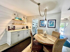 A dining area with a round table and wicker chairs, a kitchen with white cabinets, and a hanging light fixture. - Summer Special – Condo Steps to Pier & Shops (St Simons Island)