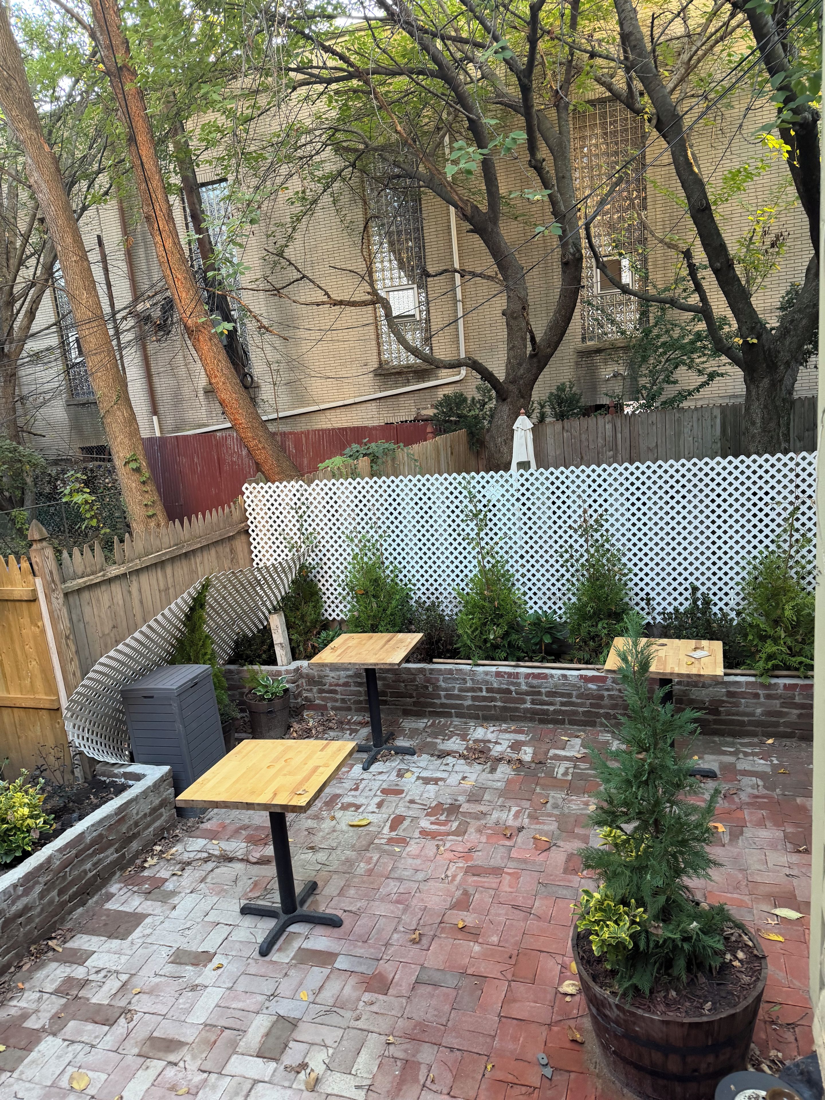 Un patio exterior con suelo de ladrillo, mesas de madera y una valla de celosía blanca.