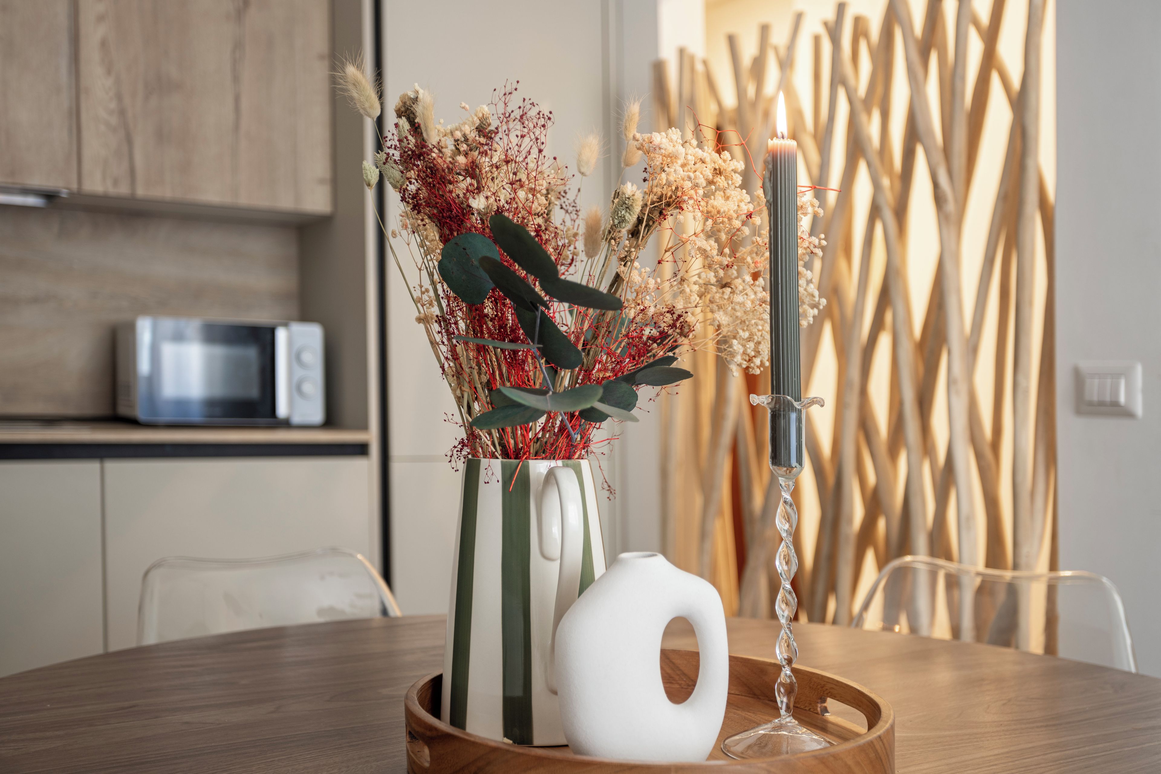 Une table à manger avec un bol en céramique blanche, un vase rayé contenant des fleurs séchées et un chandelier avec une bougie allumée.