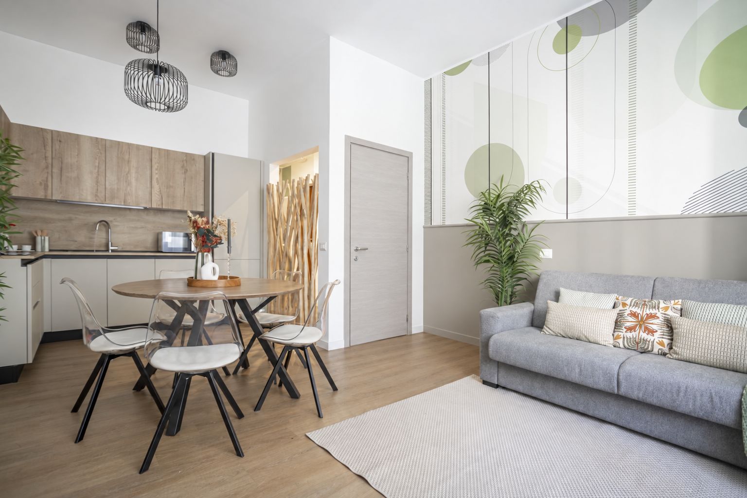 Un soggiorno moderno con un divano grigio, un tavolo da pranzo rotondo con sedie, un angolo cottura con mobili in legno e un'ampia finestra decorata con arte astratta.