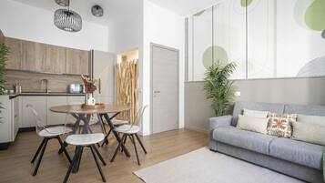 Un soggiorno moderno con un divano grigio, un tavolo da pranzo rotondo con sedie, un angolo cottura con mobili in legno e un'ampia finestra decorata con arte astratta.