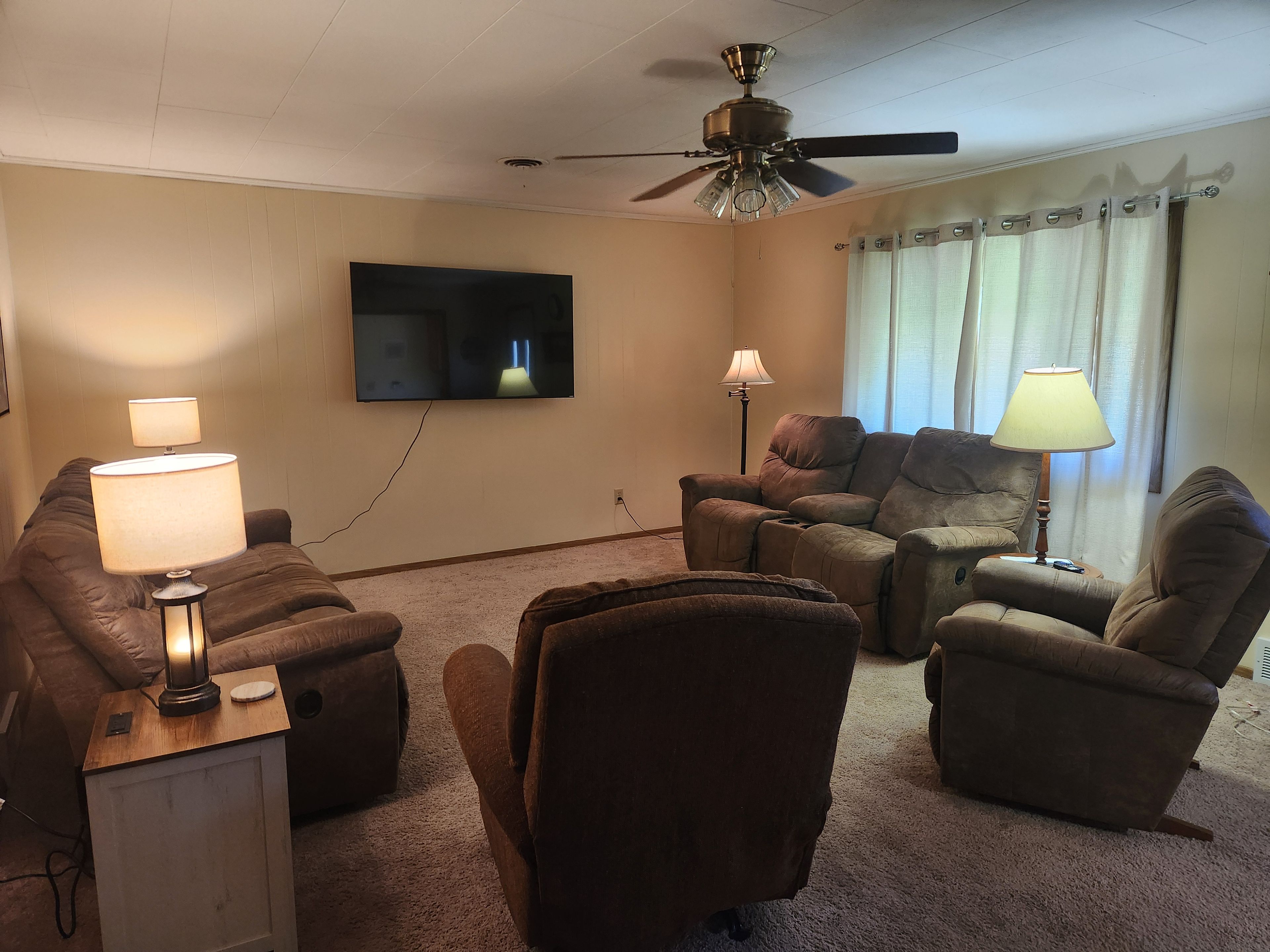 Una sala de estar con ventilador de techo, televisor de pantalla plana montado en la pared y varias opciones de asientos, incluyendo un sofá y dos sillones.