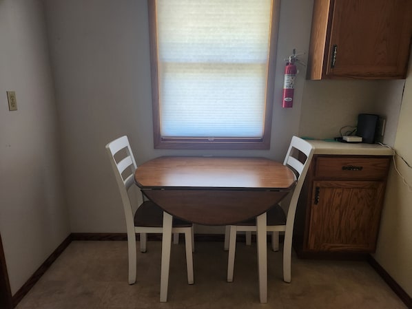 A dining area with a wooden table and two chairs, a window with blinds, a fire extinguisher, and a small wooden cabinet.