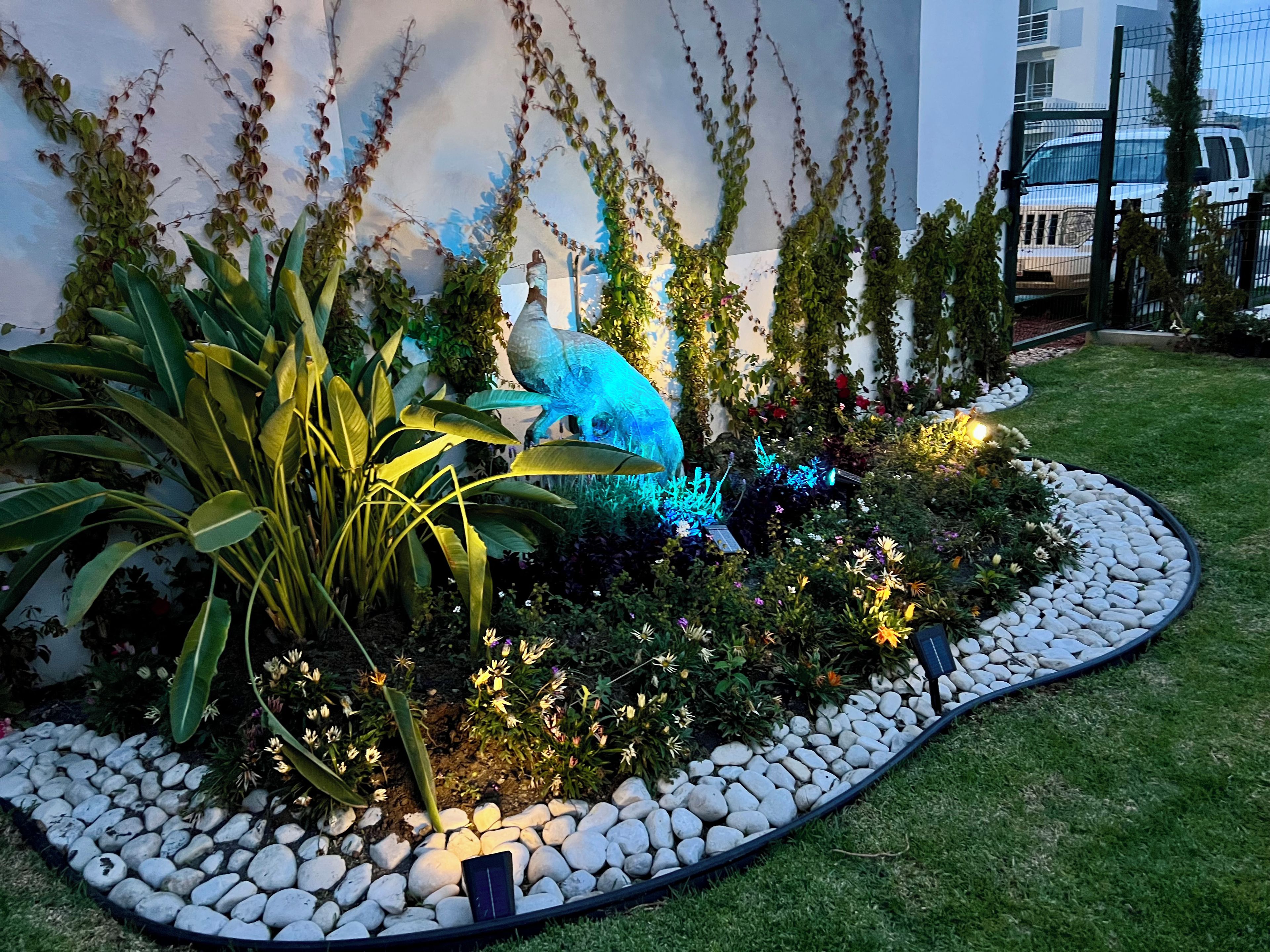 A landscaped garden with a variety of plants, a bird sculpture, and a pathway of white stones.