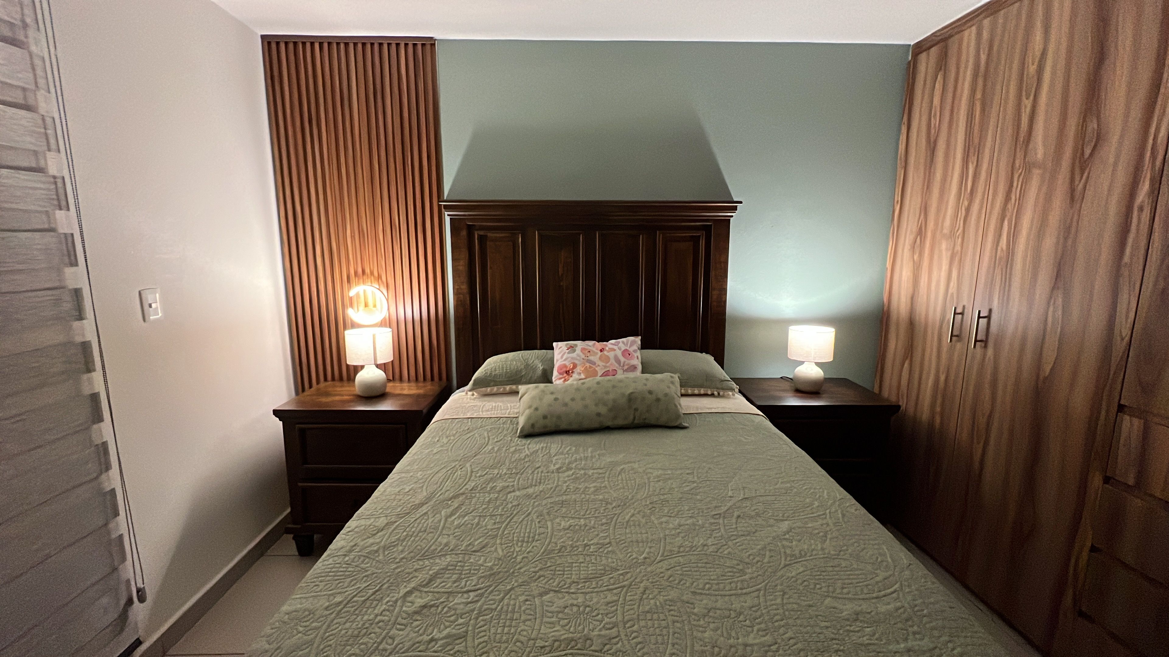 A bedroom with a bed, two bedside tables with lamps, a wooden wardrobe, and a wall with a vertical wooden panel.