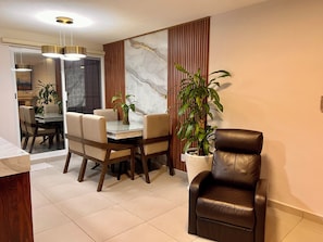 Een eethoek met een glazen tafel, houten stoelen, een plant in pot en een leren bank.