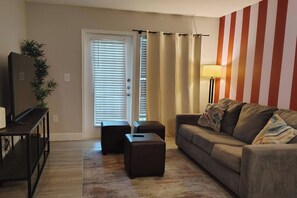 A living room with a sofa, two ottomans, a television on a stand, a lamp, and a door with curtains. - Rosie Blush 1BR 2 Beds | 5 Min Walk NRG | Near TMC (Houston)