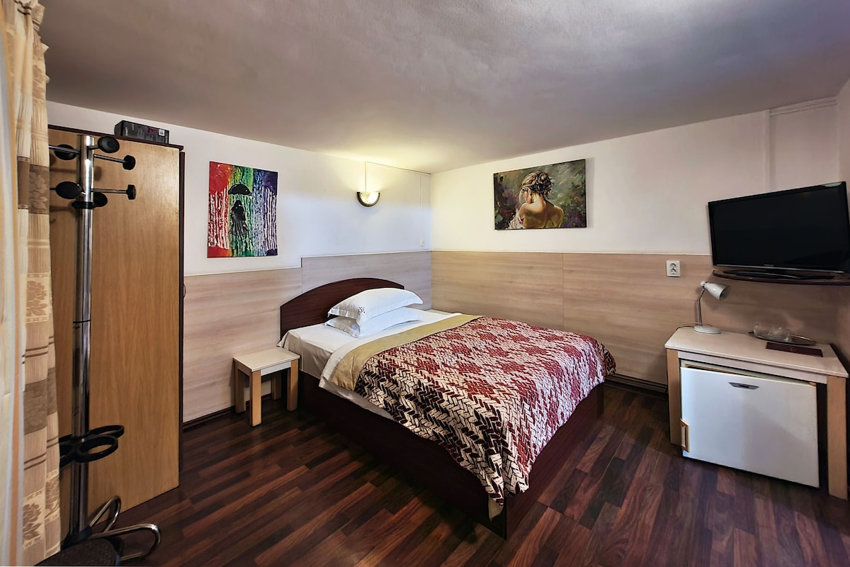 Une chambre d’hôtel comprenant un lit, une table de chevet, un petit bureau, une armoire, un téléviseur et des œuvres d’art accrochées aux murs.