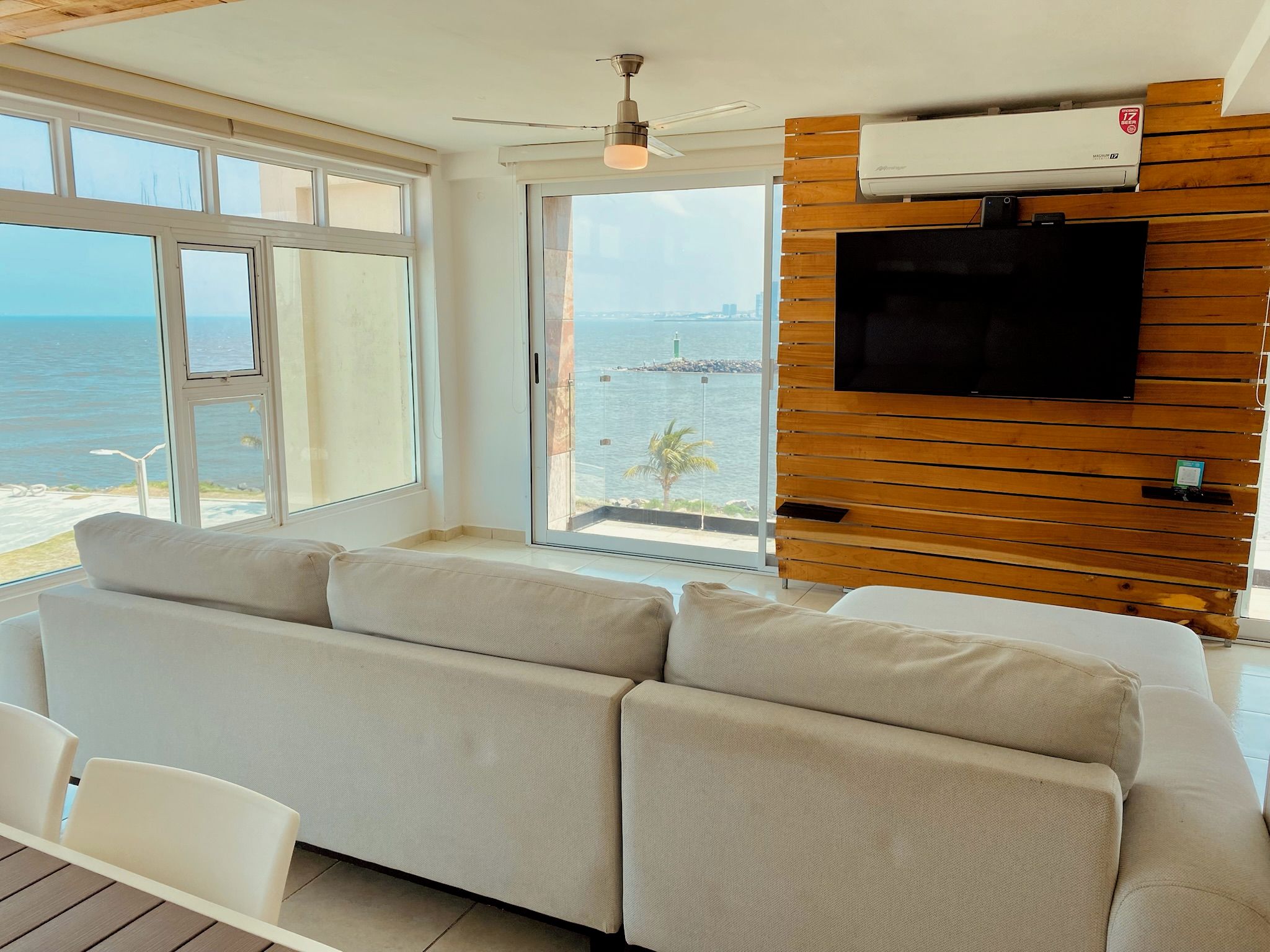 A modern living room with a sofa, a TV mounted on a wooden wall, and a view of the sea.