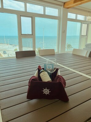 Une table avec une bouteille de vin et un verre, le tout dans un sac orné d’un motif de roue de navire, avec vue sur une plage et la mer.