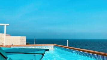 A pool with a clear view of the ocean, wooden deck, and a lounge chair.