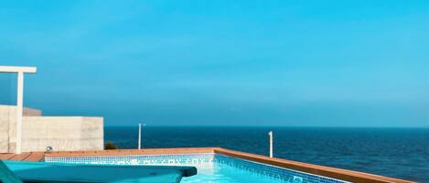 A pool with a clear view of the ocean, wooden deck, and a lounge chair.