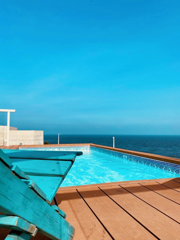 Piscina con vista despejada al océano, terraza de madera y una silla de descanso.