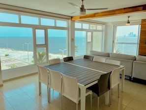 Un coin repas avec une table et des chaises, de grandes fenêtres donnant sur la mer et un ventilateur de plafond.