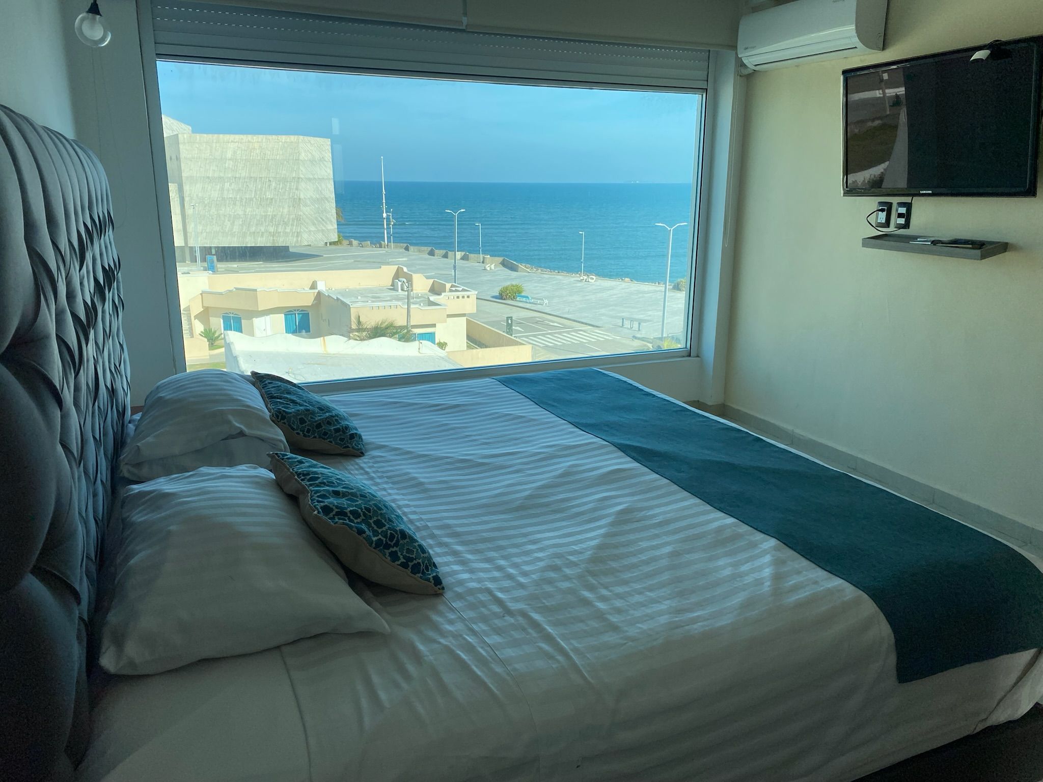 A bedroom with a large window, a bed with pillows, a flat-screen TV mounted on the wall, and a view of the sea and buildings.