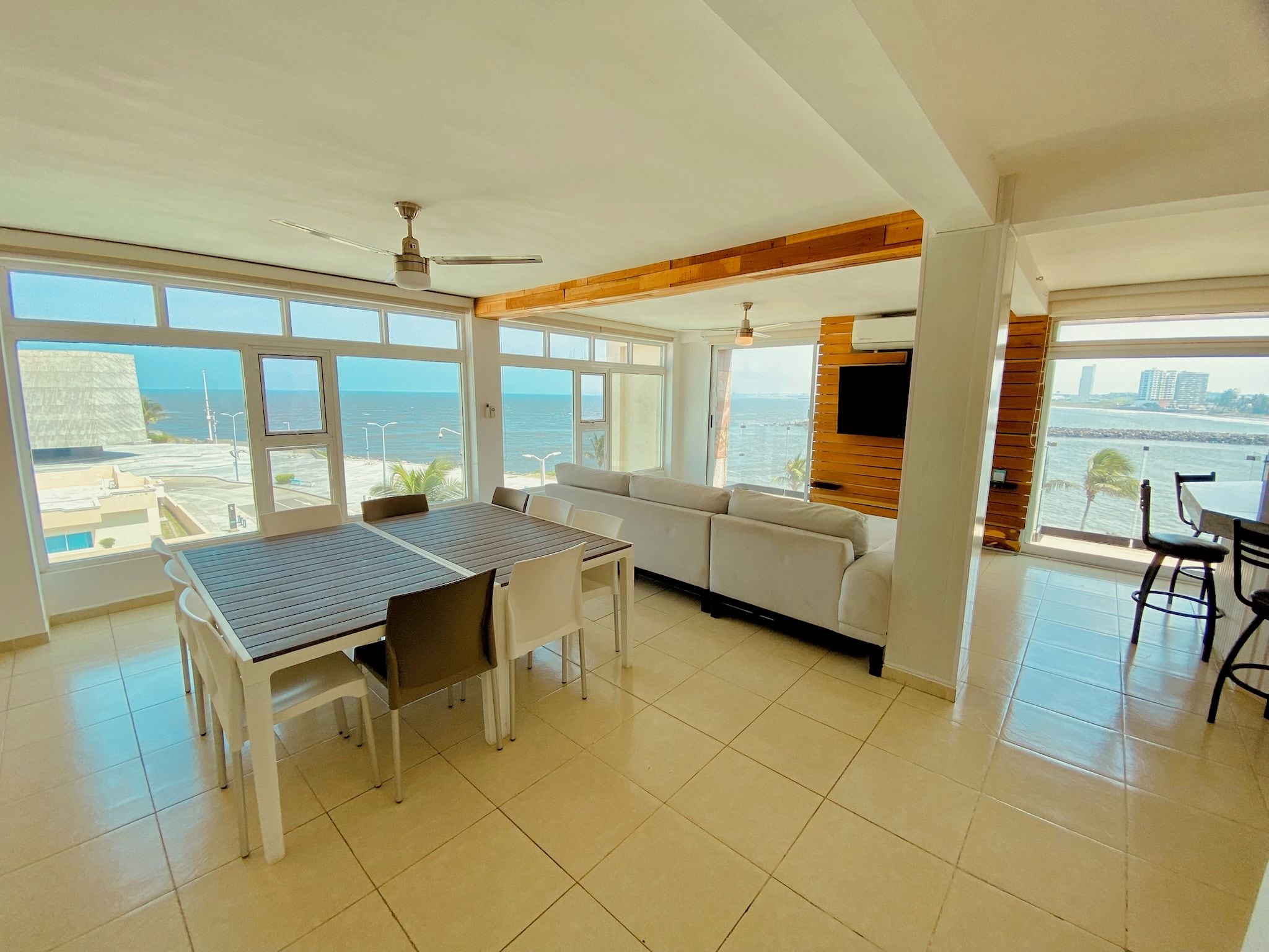 A spacious living area with a large window overlooking the sea, a dining table with chairs, a sofa, and a flat-screen TV mounted on a wooden wall.