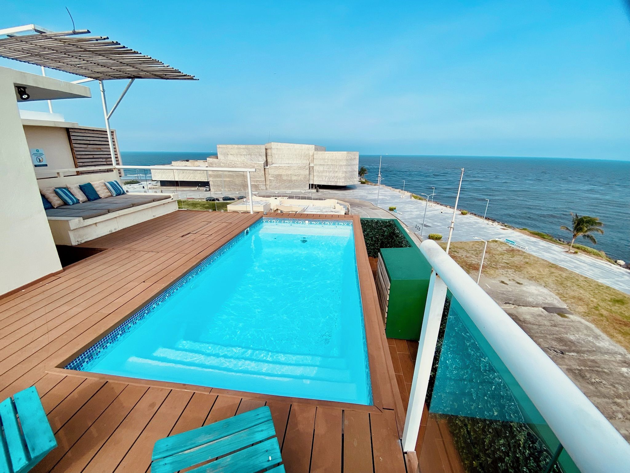 A swimming pool with a wooden deck, a lounge area with cushions, and a view of the sea.