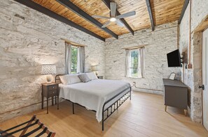 Een slaapkamer met een stenen muur, een bed, een nachtkastje, een lamp, een televisie en een houten vloer.