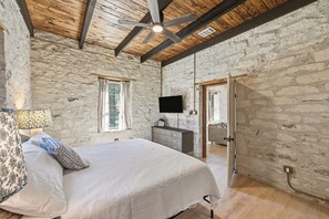 A bedroom with a bed, a dresser, a TV, and a stone wall. - Iltis Hus on Lisbon | Historic 1853 Limestone Home (Castroville)