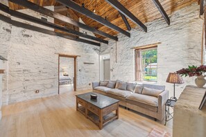 A rustic interior with exposed wooden beams, a stone wall, a sofa, a coffee table, and a window with curtains. - Iltis Hus on Lisbon | Historic 1853 Limestone Home (Castroville)