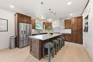 A modern kitchen with wooden cabinets, a stainless steel refrigerator, a central island with stools, pendant lighting, and a view of greenery outside. - Iltis Hus on Lisbon | Historic 1853 Limestone Home (Castroville)