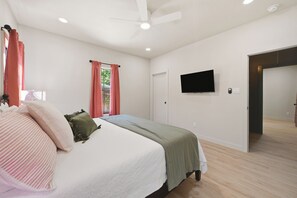 A bedroom with a bed, a flat-screen TV mounted on the wall, and a door leading to another room. - Iltis Hus on Lisbon | Historic 1853 Limestone Home (Castroville)