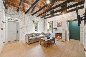 A rustic room with exposed wooden beams, a stone wall, a fireplace, and a sofa. - Iltis Hus on Lisbon | Historic 1853 Limestone Home (Castroville)