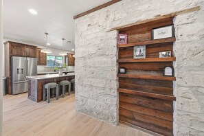 A modern kitchen with a stone accent wall, wooden shelves, and a stainless steel refrigerator. - Iltis Hus on Lisbon | Historic 1853 Limestone Home (Castroville)