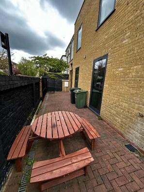 Un patio de ladrillo con una mesa y bancos de madera, una cerca negra y un edificio con ventanas.