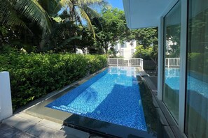 Una piscina rectangular con suelo de baldosas, rodeada de exuberante vegetación y una valla blanca.
