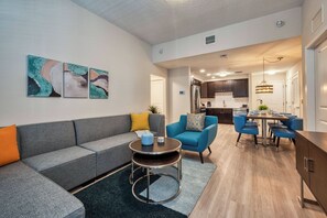 Un salon moderne avec un canapé gris, un fauteuil bleu et une table basse. On aperçoit une cuisine à l’arrière-plan, avec des meubles de couleur sombre, et un coin repas avec des chaises bleues.