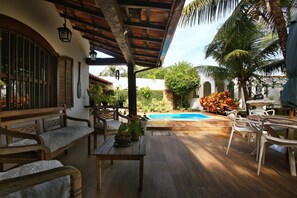 Un patio techado con mobiliario de madera, una piscina y plantas tropicales.