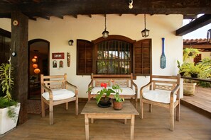 A wooden patio with two chairs and a table, a window with shutters, and a wall-mounted sign. - Casa Alto Astral em Buzios com Piscina (Búzios)