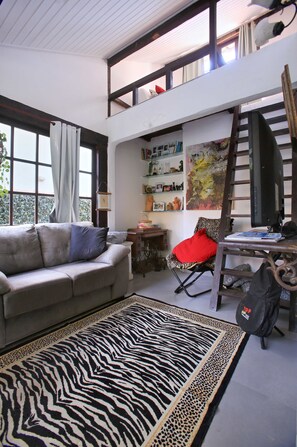 Ein Loft-Wohnraum mit hohen Decken, einer Couch, einem Leopardenfell-Teppich, einem roten Sessel und einer Leiter.