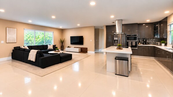 A modern kitchen with stainless steel appliances, a dining area with a white table, and a black sofa in a spacious living room with a large window. - The Quiet Luxury (Hollywood)